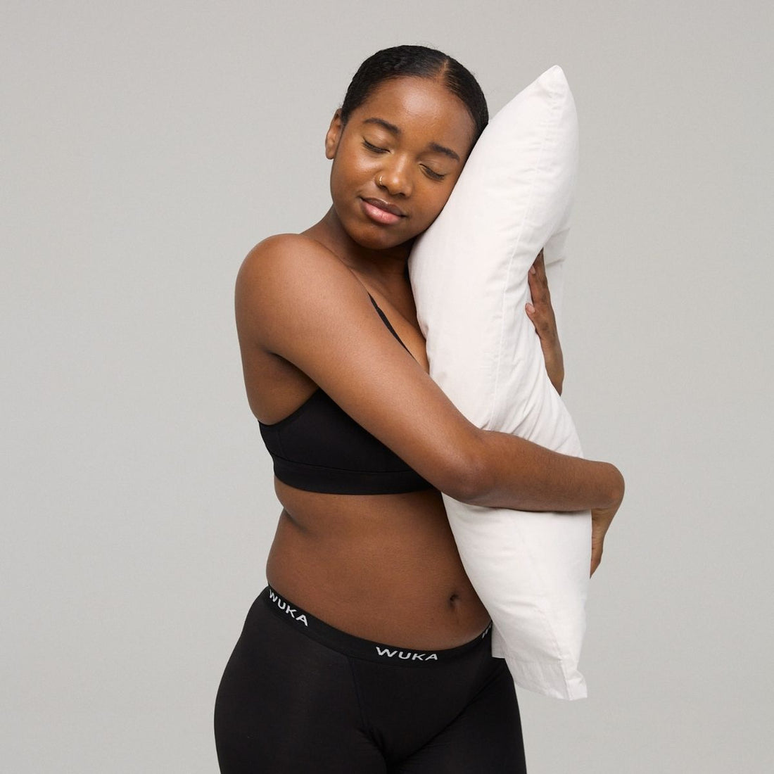 Woman in black WUKA period underwear hugs a white pillow with eyes closed, standing against a neutral background—calm, comfortable, and restful.